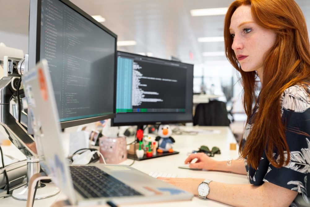 Red-haired woman sitting by a desk, coding on her computer