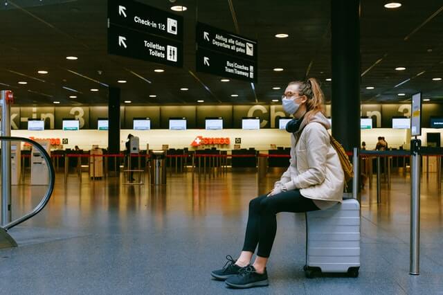 Young woman wearing a mask sits on top of her suitcase, while waiting on airport lobby