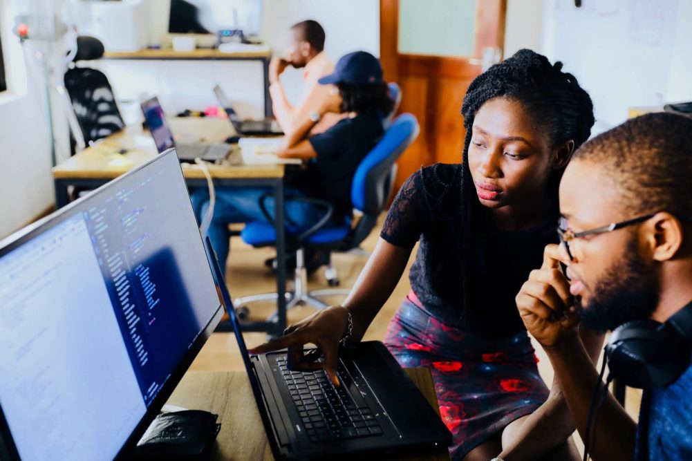 We see a room with four black software engineers. A woman is supervising a male co-worker's code. Photo by heylagostechie on Unsplash
